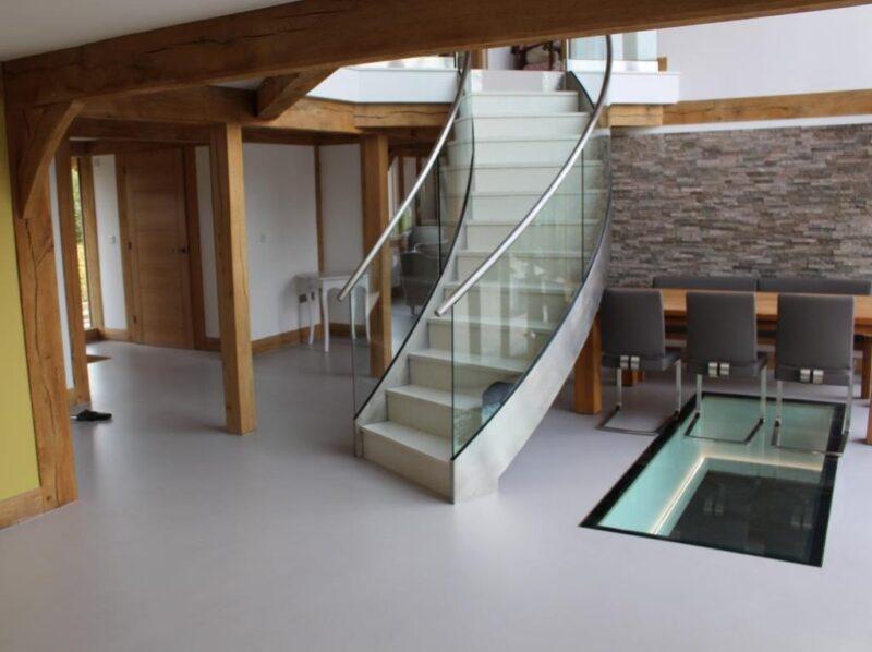Luxury Residential Resin Flooring Installation A high-end residential interior featuring a seamless light grey resin floor that extends throughout an open-plan living space, complemented by oak beams, a curved glass staircase, and a feature floor window.