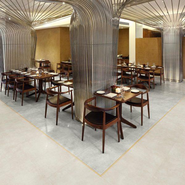 A stylish restaurant interior featuring wooden tables and chairs with black cushions, arranged neatly. The space is defined by sleek, metallic, wave-like columns and ceiling structures, creating an elegant and contemporary atmosphere. The floor is tiled with light-colored tiles accented by a gold trim, and the walls are adorned with warm beige tones.