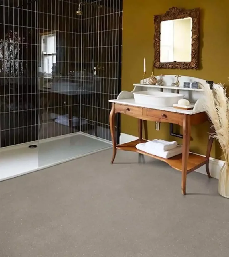 Durable LVT Flooring for Eclectic Bathrooms with Antique Vanities Light grey speckled LVT flooring in an eclectic bathroom with a wooden antique vanity, ornate mirror, and dark tiled walk-in shower.