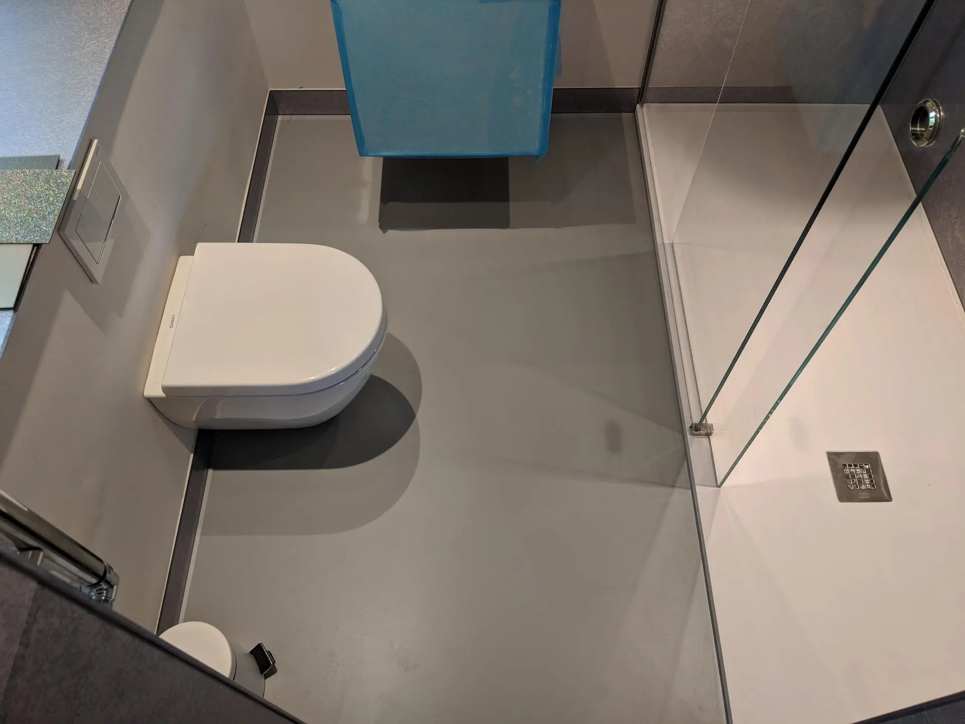 Seamless Grey Microcement Wet Room Floor for Modern Walk-In Showers Aerial view of a modern, seamless medium-grey microcement floor in a wet room with a wall-hung white toilet and a glass walk-in shower enclosure.