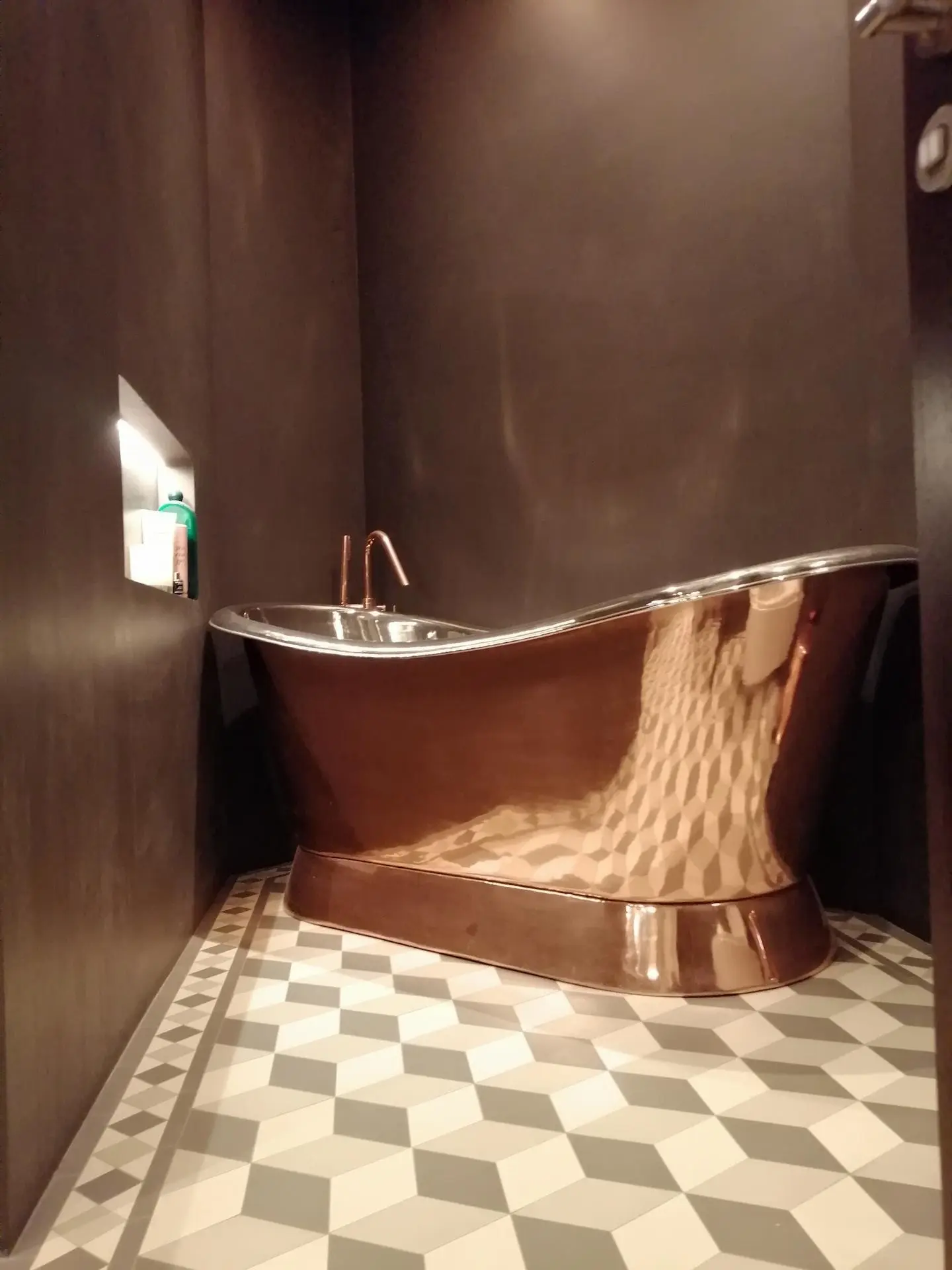 Copper Freestanding Bathtub with Seamless Dark Microcement Walls and Geometric Tiles Luxury dark bathroom featuring seamless charcoal microcement walls, a geometric patterned tiled floor, and a striking copper/rose gold freestanding roll-top bathtub.
