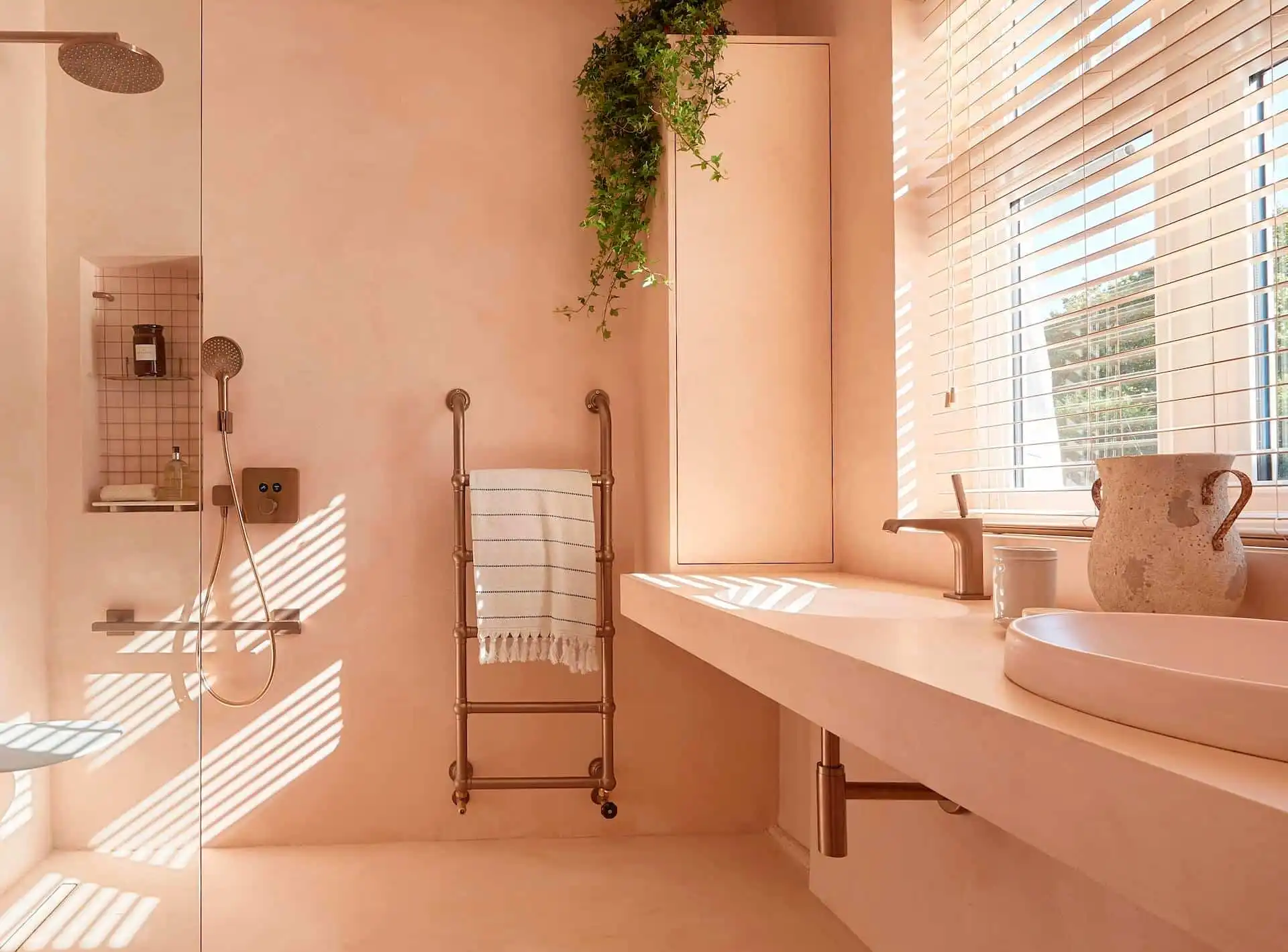 Seamless Peach Microcement Bathroom with Integrated Furniture and Wet Room Design Luxury bathroom with seamless peach microcement covering walls, floor, and integrated vanity unit, featuring a glass walk-in shower and a rustic bronze heated towel rail.
